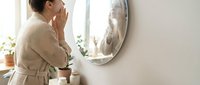 Young woman in beige robe applying skincare product while looking in bathroom mirror during morning routine. Self-care and beauty concept for wellness lifestyle.
