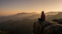 Person meditating on mountain peak at sunrise with layered hills and warm golden light creating peaceful atmosphere for wellness and mindfulness content.