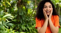 Young woman with long dark curly hair and olive skin tone wearing bright orange shirt, smiling with hand to face in lush green garden setting for lifestyle and wellness content.