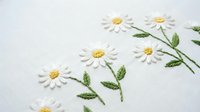 Delicate white daisy flowers with yellow centers hand embroidered on white fabric using traditional needlework techniques for crafting and textile art projects.