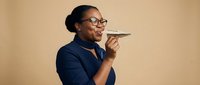 Professional African American businesswoman in navy suit speaking into smartphone on speaker mode against neutral beige background for corporate communication concepts.