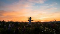 Woman in flowing dress running through wildflower meadow at golden hour sunset, arms outstretched in freedom and joy, surrounded by blooming flowers and natural landscape.
