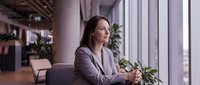 Thoughtful Caucasian businesswoman in gray blazer sitting by office window, looking away with focused expression in modern workspace.