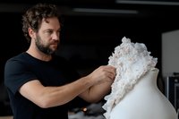 Caucasian male fashion designer with beard adjusts white lace fabric on mannequin in dark studio workspace, focused on creative dressmaking process.