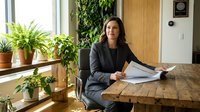 Professional businesswoman in gray suit reviewing documents at wooden desk in modern office with green plants and natural lighting for corporate presentations.