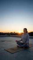 Young woman practicing yoga meditation on rooftop at sunrise with city skyline background for wellness and mindfulness lifestyle content.