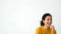 Young woman with dark wavy hair and olive complexion wearing mustard yellow ribbed sweater, smiling while touching face against white background for portrait and lifestyle use.