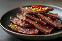 Spicy glazed beef jerky strips garnished with sesame seeds and green onions on dark plate for snack food marketing and culinary photography.