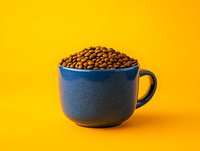 Fresh roasted coffee beans overflowing from navy blue ceramic cup against vibrant yellow background, minimalist composition for food and beverage marketing.