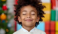 Happy mixed-race child with curly hair and closed eyes smiling peacefully in white sweater against colorful Christmas background with tree and gifts.