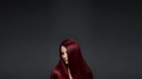 Young woman with long burgundy hair, fair complexion, looking downward against dark gray background, suitable for beauty, fashion, and salon marketing materials.