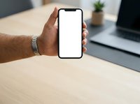 Man holding smartphone with blank white screen at wooden desk with laptop and plant, displaying mobile device for app presentation or website mockup.