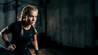 Young athletic woman with blonde braided hair, Caucasian ethnicity, wearing black sportswear, running indoors in industrial gym setting with dramatic lighting and concrete walls.
