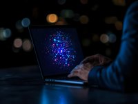 Person working on laptop with glowing blue and purple digital particle visualization display against dark background with bokeh lights.