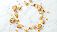 Scattered golden cereal pieces and crumbs arranged in circular pattern on white marble surface, overhead view for food styling, breakfast advertising, and culinary design projects.