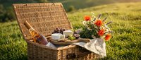 Wicker picnic basket with food, wine, and fresh flowers on green grass for outdoor dining and summer leisure activities.