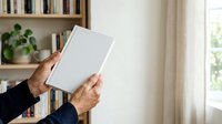 Senior adult with dark skin tone holding blank white book cover in home library setting with shelves and natural window light, suitable for publishing, education, and lifestyle marketing.