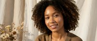 Young African American woman with curly hair smiling warmly in cozy brown sweater by window with dried flowers for lifestyle and portrait content.