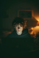 Young person with curly hair illuminated by warm light in dark room, face glowing from screen, wearing black hoodie in moody atmospheric setting.