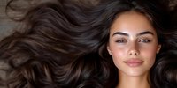 Young woman with flowing dark hair and natural makeup lying down, showcasing healthy glowing skin and radiant smile against neutral background. Perfect for beauty and haircare advertising.