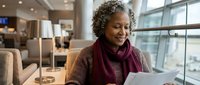 Mature African American woman with curly gray hair reading documents in modern office lobby wearing burgundy scarf and brown sweater by large windows.