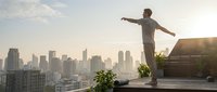 Young man standing on urban rooftop with arms outstretched overlooking city skyline at sunset, expressing freedom and achievement in metropolitan setting.