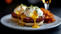 Eggs Benedict with crispy bacon on English muffin, dripping hollandaise sauce and fresh herbs. Served with golden fried potatoes on white plate against dark background.
