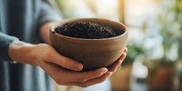 Person holding wooden bowl with rich dark soil for gardening or planting, with soft natural light and green plants in background.