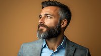 Middle-aged Caucasian businessman with gray beard and styled hair wearing blue shirt and gray suit jacket gazes confidently against warm brown background.