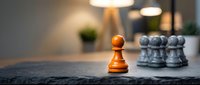 Golden pawn standing alone facing silver pawns on stone surface with warm office lighting, representing leadership, strategy, competition and business success.