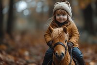 Young child wearing knit beanie and brown coat riding pony in autumn forest setting with fallen leaves and natural bokeh background, capturing childhood joy.