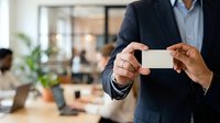 Businessman in navy blazer and light blue shirt holding blank white business card in modern office environment with blurred colleagues and plants in background.
