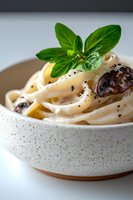 Creamy fettuccine pasta with black olives and fresh basil garnish in white ceramic bowl, served with black pepper seasoning on light background.