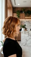 Young Caucasian woman with wavy auburn bob haircut in profile view wearing black top in modern kitchen interior with marble countertops and wooden cabinets.