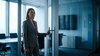 Professional Asian businesswoman in gray suit opening glass door in modern corporate office with city view and conference room background.