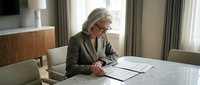 Senior woman with gray hair and glasses reviewing architectural blueprints and documents at a marble table in a modern office space with natural light from windows.
