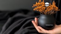 Black ceramic pot with dried orange flowers and decorative skull held in feminine hand against dark background, moody Halloween party decor setup.