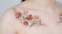 Delicate floral collarbone tattoo on a young woman's skin, featuring watercolor orange and yellow wildflowers with green leaves.