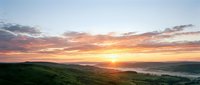 Dramatic sunrise over rolling green hills and misty valley with golden sun breaking through orange and pink clouds, scenic landscape for nature and travel projects.