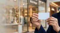 Woman holding blank white business card in front of luxury retail storefront window display for branding, marketing, and corporate identity mockup purposes.