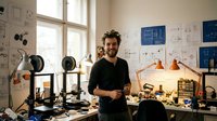 Young male engineer smiling in modern workshop with 3D printers and technical blueprints on walls. Innovation and technology development workspace.