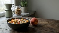 Healthy breakfast cereal with fresh fruit in black bowl on wooden table, morning meal setup with coffee maker and potted plant.