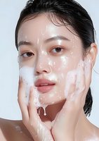 Young Asian woman with glowing skin applying facial cleanser foam, demonstrating skincare routine against white background. Closeup beauty portrait shows foam texture.