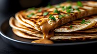 Stack of homemade crepes with caramel sauce dripping down sides, garnished with fresh green herbs on dark plate, close up view with selective focus.