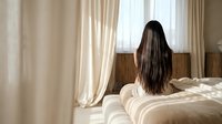Young Asian woman with long dark hair sitting on bed in bright bedroom with natural sunlight streaming through curtains creating peaceful morning atmosphere.