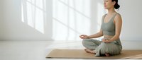 Young Asian woman practicing meditation in lotus position on yoga mat in bright minimalist room with natural sunlight shadows on wall.