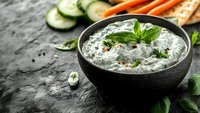 Fresh tzatziki sauce with cucumber, herbs and garlic served in black bowl on dark stone background with vegetables and bread for dipping.