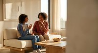 Two young African American women having friendly conversation while sitting on modern beige sofa in bright living room with natural sunlight streaming through windows.