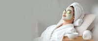 Young Asian woman relaxing in spa with green clay face mask and cucumber slices, wearing white robe and towel turban on head.