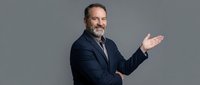 Professional middle-aged Caucasian businessman in dark suit presenting with open hand gesture against gray background for corporate marketing materials.